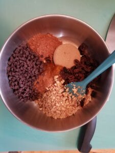Metal bowl containing a teal spatula and unmixed filling ingredients: brown sugar, raisins, chopped walnuts, apricot jam, cinnamon, mini chocolate chips, and cocoa powder.