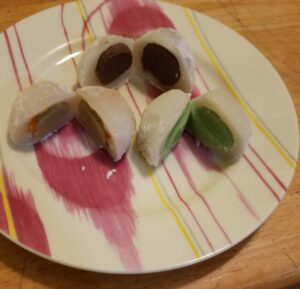Three daifuku mochi, cut in half to show centers, arranged on a pink-swirled plate. From top: chocolate center, green honeydew center, and tan mango center with mango bits.