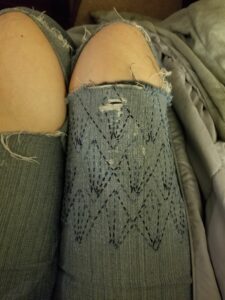 Front thigh panel of a pair of ripped blue jeans, decorated with sashiko-style stitching of variegated blue tones in an art-deco diamond fan pattern, photographed while worn.