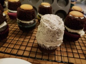 Several chocolate cupcakes, sandwiched together with frosting, resting on a grid-style black cooling rack. In the foreground, one such stack is crumb-coated with white frosting. In the background, several other stacks, each topped with a vanilla wafer cookie, await their crumb coat.