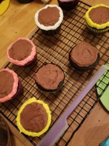 Several chocolate cupcakes on a black grid-style cooling rack, all frosted in chocolate, some with rings of colored icing around their edges.