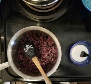 Pot of mixed blueberries and sugar on a stove, with a pirate-printed rubber spatula resting in the pot. A small glass bowl of cornstarch solution waits at right, with a fork in it to stir.