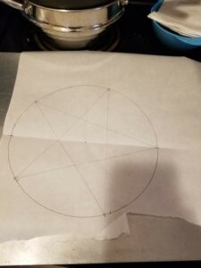 A cookie sheet holding a piece of parchment paper, with a large circle drawn on it in pencil and a five-pointed star drawn inscribed in the circle.