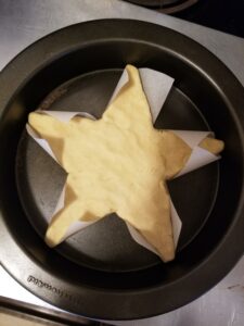 Round cake pan with a cookie-dough star in it, unbaked, with parchment paper visible around the points of the star.