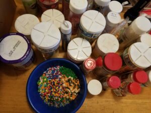 A blue bowl containing multiple kinds of decorative sprinkles, surrounded on a tabletop by many bottles of sprinkles.