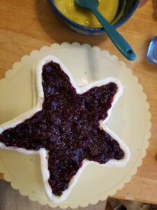 Star-shaped cake on a rotating cake plate, outlined with white icing and topped with blueberry filling.