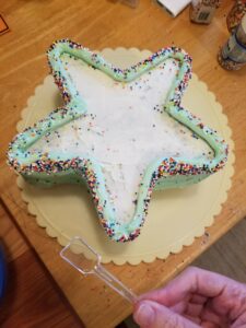 Star-shaped cake on a rotating cake plate, top lightly frosted in white, sides frosted in light green, and top edges heavily outlined in light green, with multicolor tiny round sprinkles applied around the top outer edge. A hand in the foreground is holding up a small clear plastic squared-off spoon/spatula.