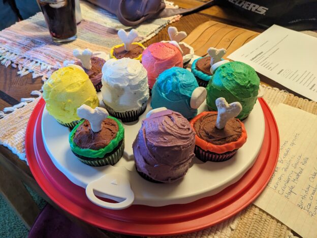 A plate of cupcakes decorated like Among Us crewmates, some double-height and alive with marshmallow faceplates and others dead with candy bones sticking up from their single-height corpses.