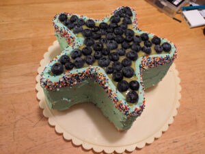 Star-shaped cake on a rotating cake plate, sides frosted in light green, top outer edge covered in multicolored sprinkles, center top filled with lime curd and topped with fresh blueberries.