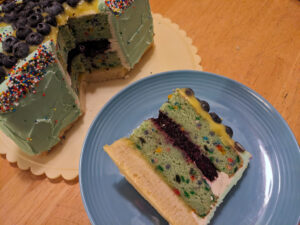 Slice of cake on a blue plate, showing a bottom layer of shortbread and two layers of green cake studded with sprinkles, with blueberry filling in the middle, frosting on the outer edge, and lime curd and fresh blueberries on top. The rest of the cake is visible in the background.