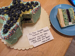 Slice of cake on a blue plate beside the rest of the cake on a rotating cake plate, showing a bottom layer of shortbread and two layers of green cake studded with sprinkles, with blueberry filling in the middle, light green frosting on the outer edges, and lime curd and fresh blueberries on top. An index card with the bottom left corner jaggedly missing rests on the cake plate, with the message: "To the shark known as Grace: Please enjoy this cake and stop eating my raft. -The Mgmt."