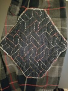 Background: black, gray, and red plaid fabric. Foreground: navy blue corduroy fabric stitched around the edge and in an all-over pattern of linked ropes with variegated red-and-gray thread.