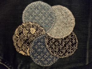 Patch made of five overlapping cloth circles in various shades of blue, arranged in a flower shape, with cross shapes stitched all over.