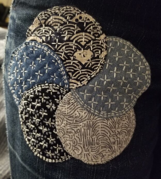 Patch made of five overlapping circles of cloth, in various shades and patterns of blue, arranged in a flower shape and stitched all over with off-white thread, on a pair of dark blue jeans.